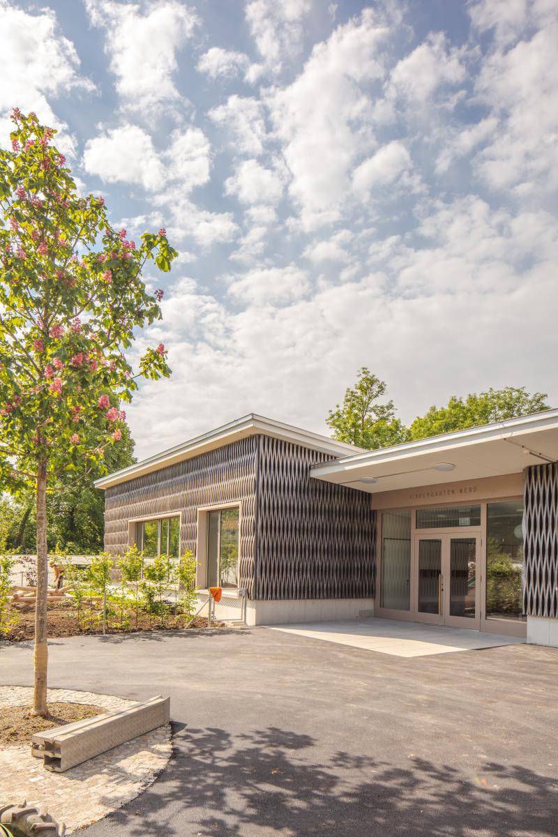 Kindergarten Werd in Adliswil mit dunkler Holzfassade, großflächigen Verglasungen und vorgelagerter Terrasse unter wolkenbedecktem Himmel.