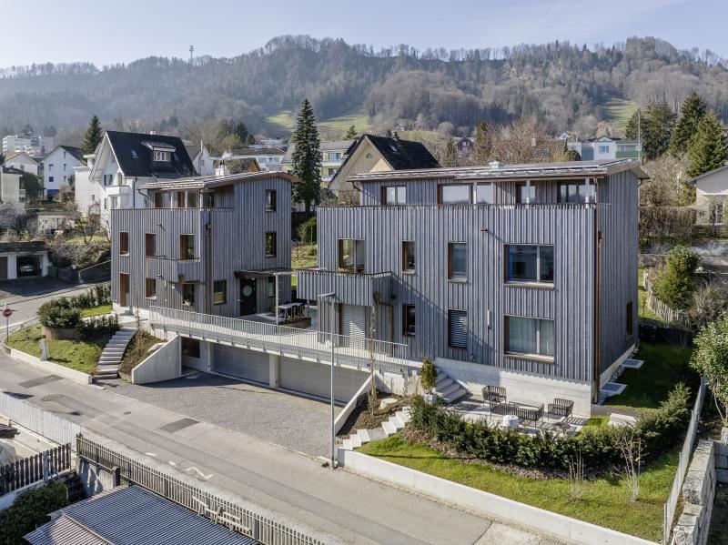 Modernes dreigeschossiges Mehrfamilienhaus mit Holzfassade in Adliswil, umgeben von Wohngebäuden und bewaldeten Hügeln.