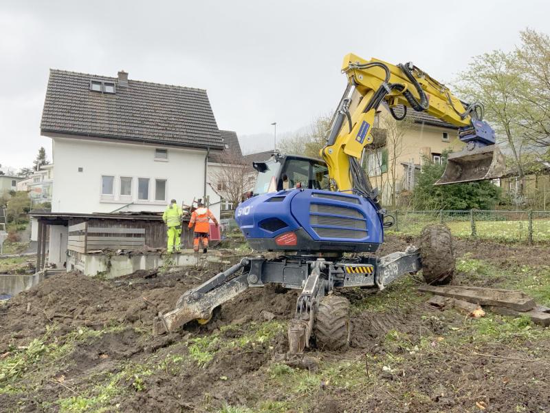 Baggerschaufel an einer Baustelle zweier Mehrfamilienhäuser in Adliswil mit Arbeitern und bestehenden Wohnhäusern im Hintergrund.