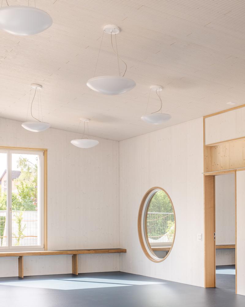 Heller Kindergartenraum mit weißer Decke, hängenden Leuchten, Holzregalen und rundem Fenster.