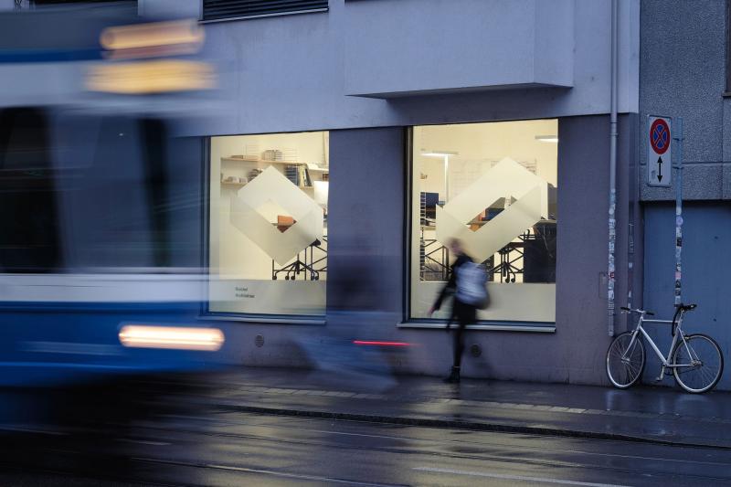 Ein vorbeifahrender Bus und ein Fußgänger vor zwei beleuchteten Schaufenstern eines modernen Bürogebäudes in der Abenddämmerung.