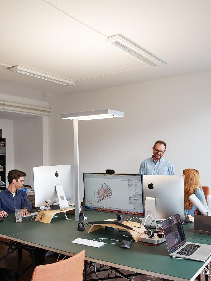 Vier Personen arbeiten an Computern und an Entwürfen an einem grünen Schreibtisch in einem Büro.