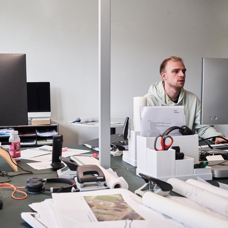 Ein Mitarbeiter sitzt an einem unaufgeräumten Schreibtisch in einem Büro und blickt auf seinen Monitor.