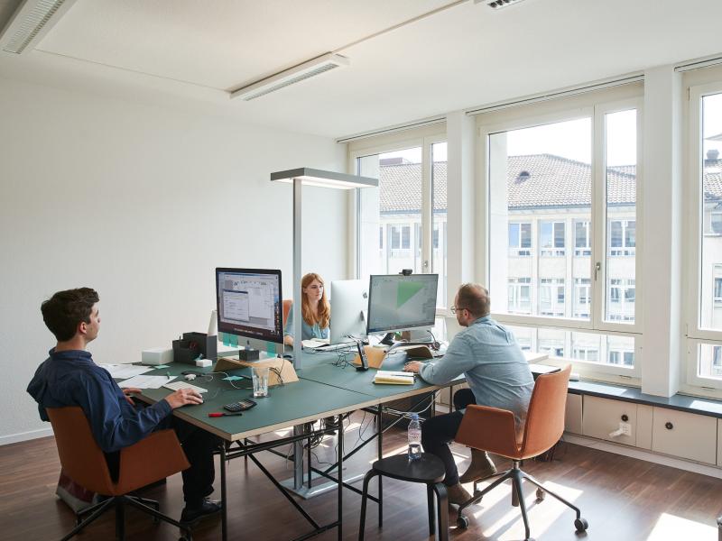 Vier Mitarbeiter arbeiten an Schreibtischen mit Computern in einem hellen Büroraum mit großen Fenstern.