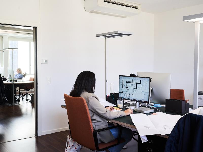 Eine Mitarbeiterin sitzt an einem Schreibtisch vor einem Monitor mit Architekturplänen in einem modernen Büro.