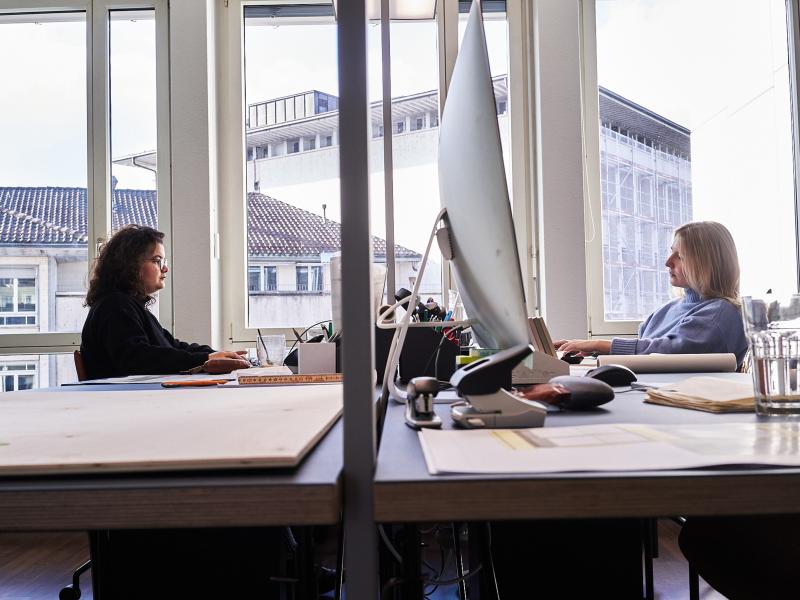 Zwei Mitarbeiterinnen sitzen sich an einem Schreibtisch in einem modernen Büro mit großen Fenstern gegenüber.
