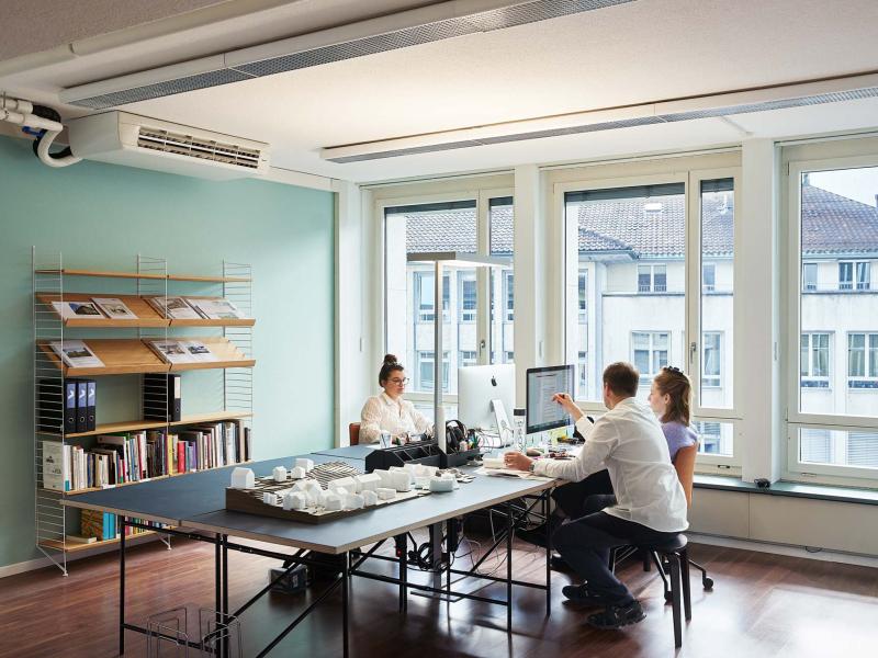 Vier Personen arbeiten an einem Tisch in einem hellen Büroraum mit türkiser Wand und großen Fenstern.