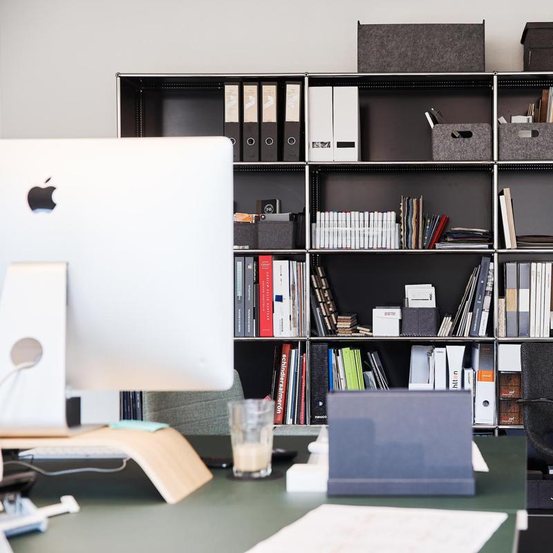 Modernes Büro mit weißem Apple-Laptop, Schreibtisch und schwarzem Wandregal gefüllt mit Büchern und Ordnern.