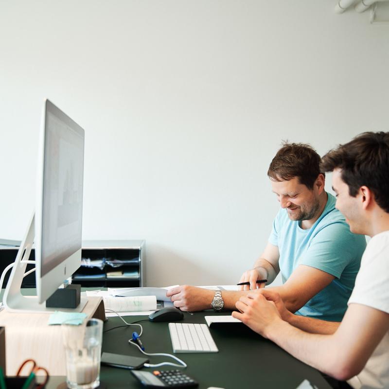 Ein Mitarbeiter und Hannes Reichel sitzen an einem Schreibtisch vor einem Monitor und besprechen gemeinsam Dokumente.