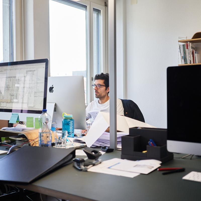 Ein Mitarbeiter sitzt an einem Schreibtisch in einem hellen Büro mit Computern und Unterlagen.