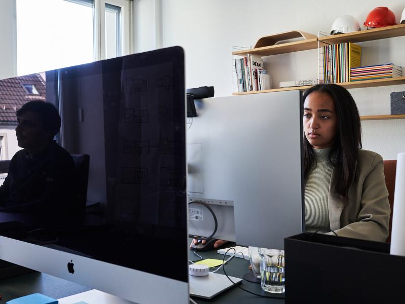 Eine Mitarbeiterin arbeitet am Computer in einem hellen Büro mit Regalen im Hintergrund.