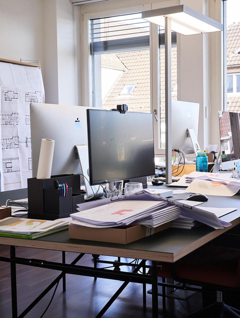 Schreibtisch des Architekturbüros Reichel in Zürich mit Computer, Bauplänen und Materialien vor einem Fenster.