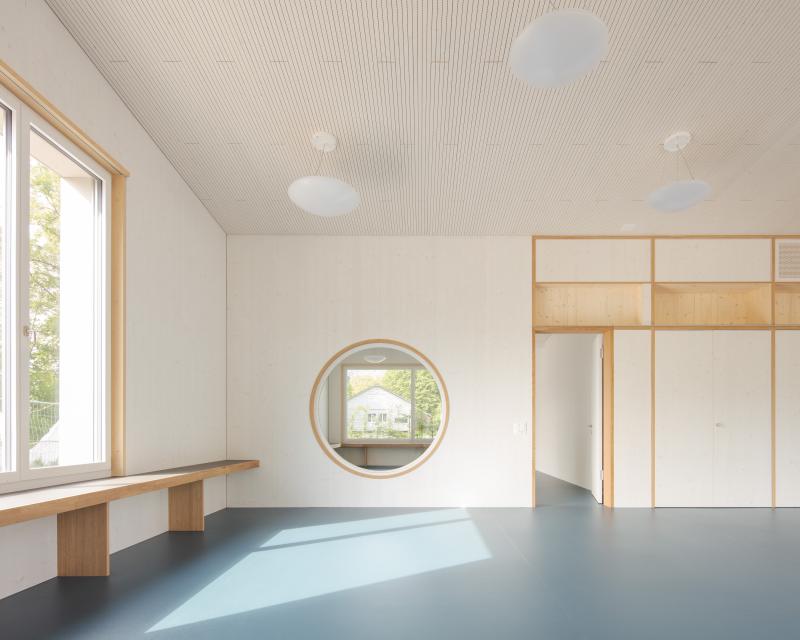 Heller Innenraum des Doppelkindergartens Werd in Adliswil mit Holzverkleidung und rundem Fenster.